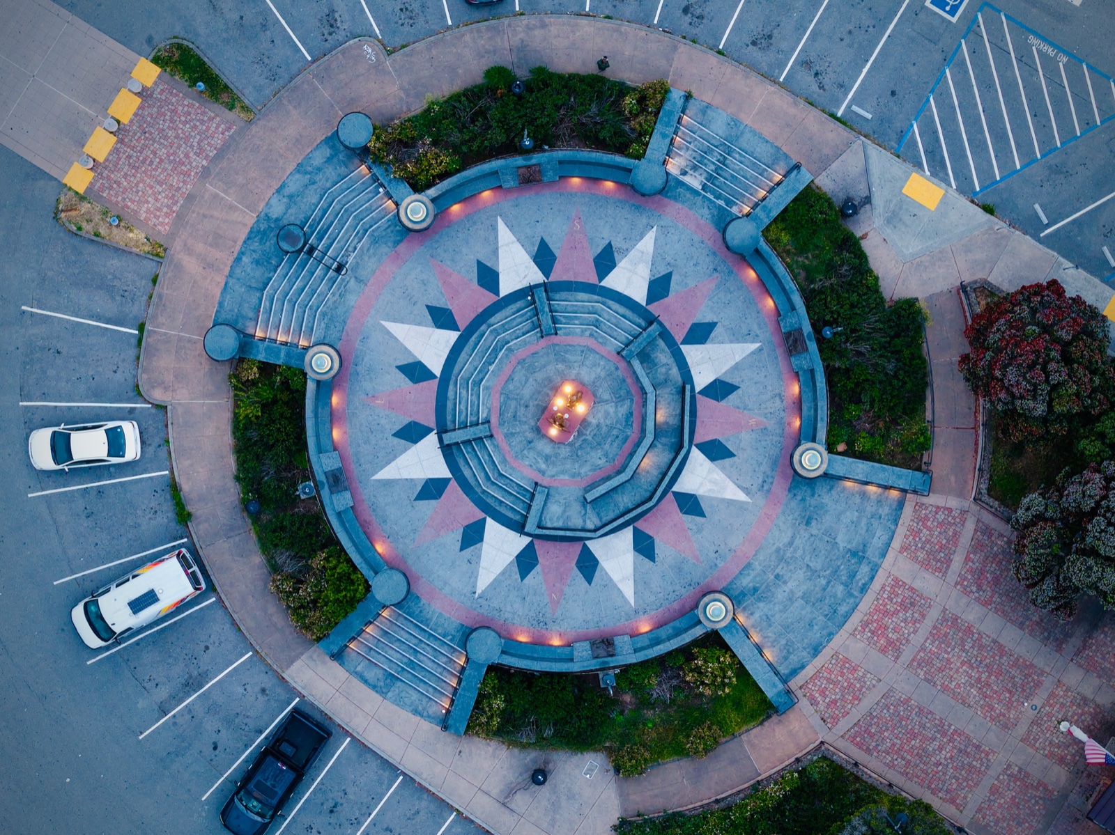 Drone photograph looking straight down on the compass plaza at Golden Gate Bridge Vista Point, with a central light surrounded by radial points, curved steps, paths, and parking areas.