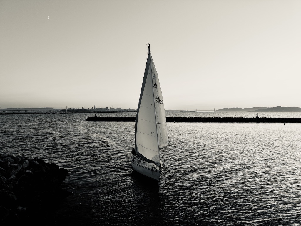 Black-and-white photograph of a sailboat near the Berkeley Yacht Club at dusk, with a crescent moon above the mast and the San Francisco skyline in the distance.