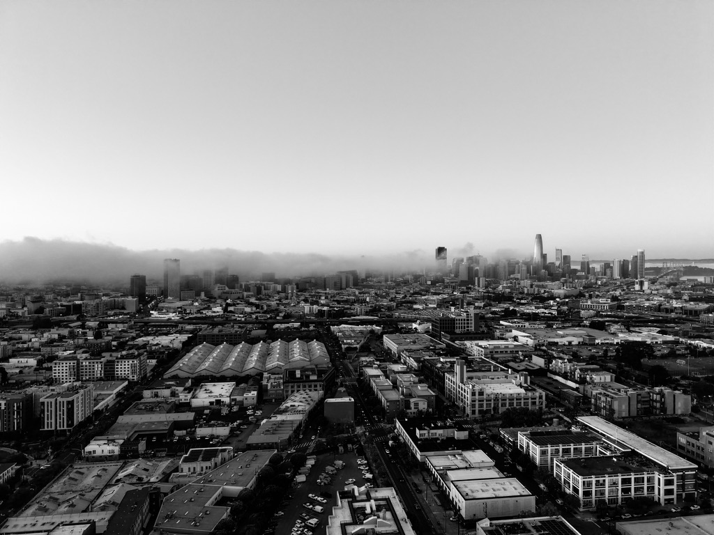 Black-and-white drone photograph of San Francisco partly obscured by fog, with the city emerging through atmospheric layers.