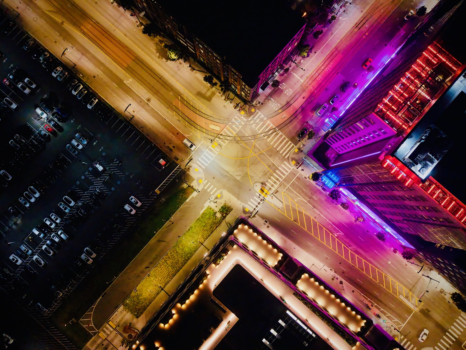Nighttime drone photograph looking straight down on a brightly lit Mission Bay intersection, with crossing routes, rail lines, moving cars, and contrasting amber, red, blue, and magenta light.